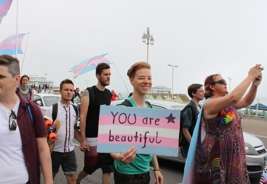 Menschen laufen demonstrierend über eine Brücke. Eine der Personen hält ein selbstgebasteltes Plakat in der Hand. Das Plakat ist hellblauen, rosa und weißen Streifen angemalt; davor steht in schwarzer Schrift: YOU are beautiful.
