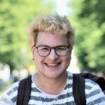 Foto von Eno, lachend mit Brille und Rucksack vor verschwommenen hellgrünen Bäumen